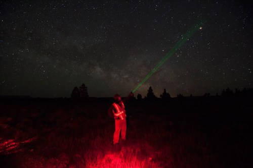 A person in a reflective vest stands in a dark field, pointing a green laser at the starry night sky.