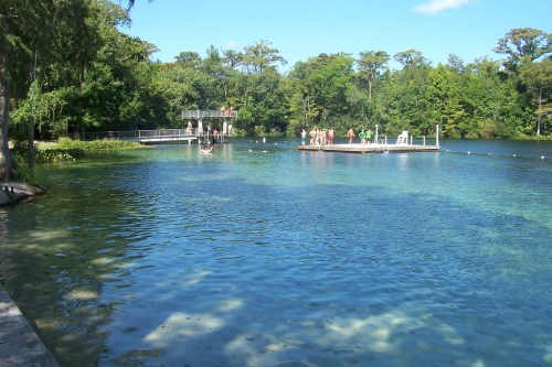 A clear blue lake with people on a dock and surrounding greenery under a bright blue sky.