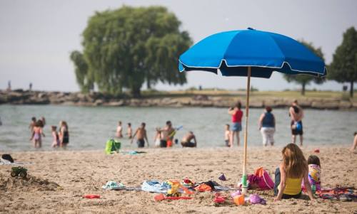 A child sits under a blue umbrella on a sandy beach, with people swimming and playing in the background.