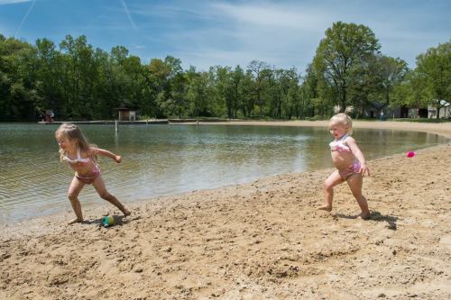 Two young children play on a sandy beach by a calm lake, enjoying a sunny day outdoors.