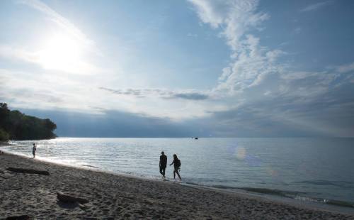 Two people walk along a sandy beach at sunset, with calm waters and a cloudy sky in the background.