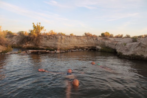 Four people float in a natural hot spring surrounded by rocky terrain and sparse vegetation under a clear sky.