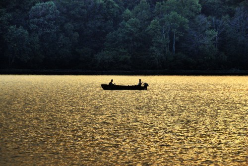 Two people in a small boat on a shimmering lake at sunset, surrounded by lush trees.