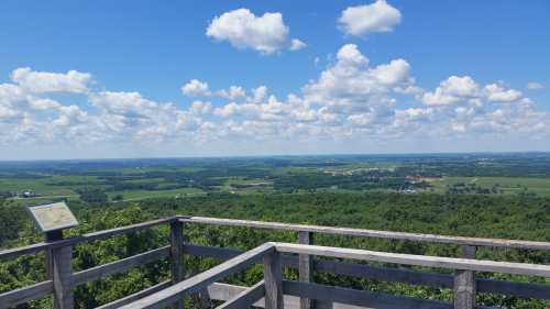 A scenic view from a lookout point, featuring lush green fields and a blue sky with fluffy white clouds.