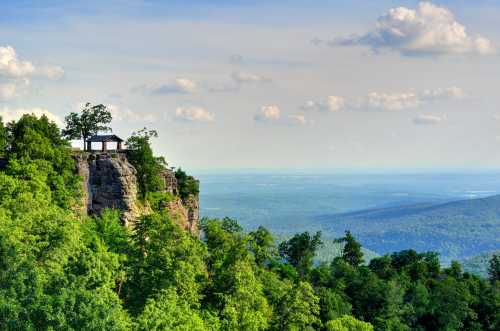 A scenic view from a cliff with a gazebo, surrounded by lush greenery and distant mountains under a blue sky.