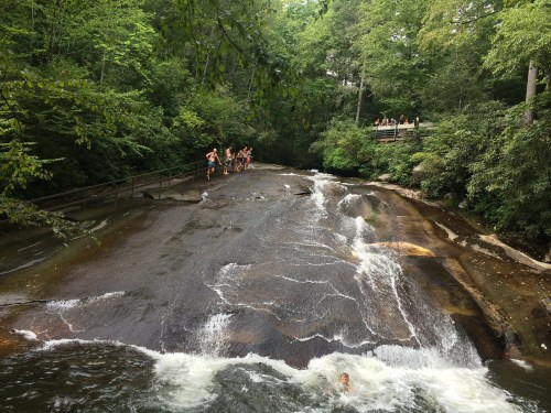 A rocky riverbed with people playing in the water, surrounded by lush green trees and a wooden observation deck above.