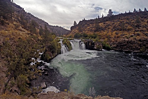 A scenic view of a waterfall cascading into a river, surrounded by rocky cliffs and sparse vegetation under a cloudy sky.