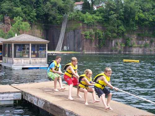 Four children in life jackets pull on a rope while standing on a dock by a lake, with a cabin and trees in the background.