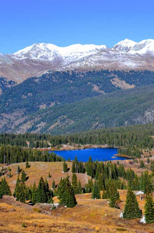 A serene mountain landscape featuring a blue lake surrounded by evergreen trees and snow-capped peaks in the background.