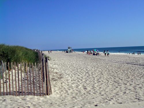 A sandy beach with people enjoying the sun, a lifeguard tower, and grassy dunes under a clear blue sky.