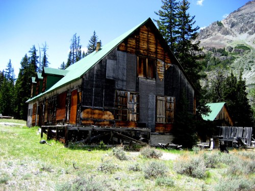 Abandoned wooden building with a green roof, surrounded by trees and mountains under a clear blue sky.