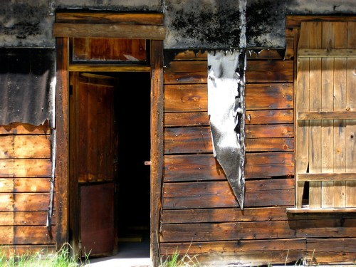 A weathered wooden building with a partially open door, showing signs of fire damage and peeling paint.