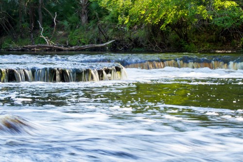 A serene river scene with gentle waterfalls and lush greenery along the banks, reflecting sunlight on the water.