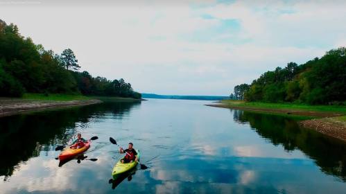 Two kayakers paddle on a calm lake surrounded by trees and a cloudy sky.