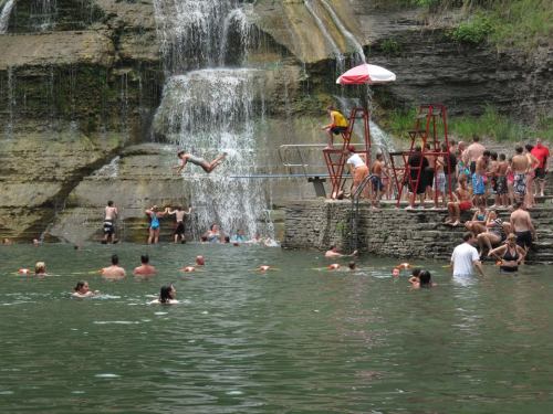 A busy swimming area with people enjoying a waterfall, diving from a platform, and swimming in a natural pool.