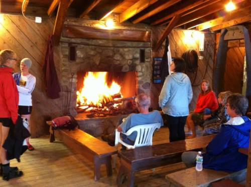 A cozy indoor space with a large fireplace, where people gather on benches, some chatting and others relaxing.