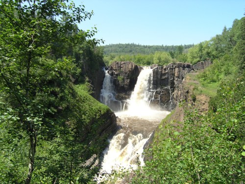 A scenic waterfall cascading over rocky cliffs, surrounded by lush greenery and a clear blue sky.