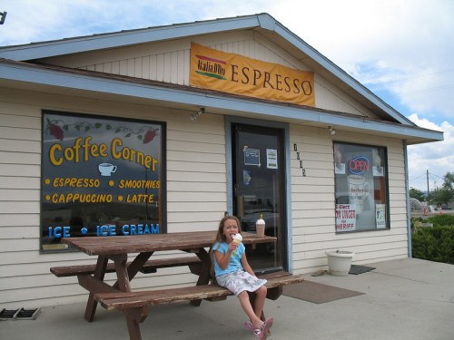 A young girl enjoys ice cream outside a coffee shop with a sign that reads "Espresso" and "Coffee Corner."