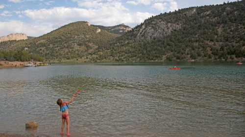 A child fishing by a lake surrounded by mountains and trees on a sunny day.