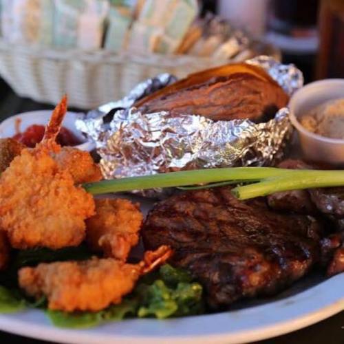 A plate of grilled shrimp, steak, and a baked potato, served with a side of dipping sauce and garnished with green onions.