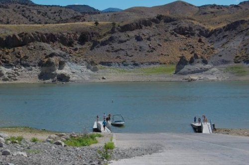 A serene lake surrounded by dry hills, with a boat ramp and a few people near the water's edge.