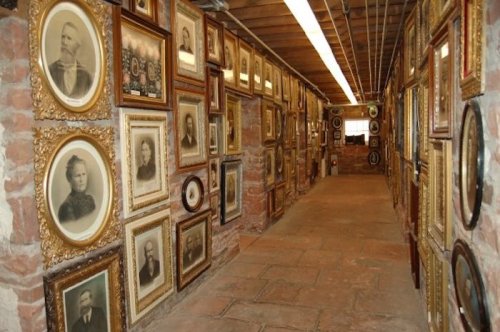A narrow hallway lined with vintage framed portraits on brick walls, illuminated by overhead lights.