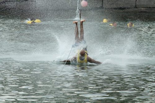A person falls into the water while being pulled by a cable, creating a splash, with others in yellow life jackets nearby.