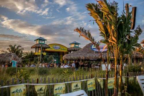 Tropical beachside restaurant with thatched roofs, palm trees, and a vibrant sunset sky in the background.