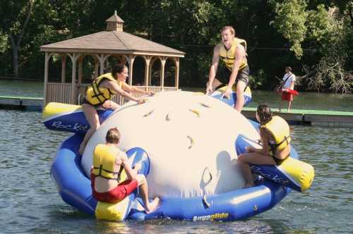 A group of people in life jackets play on a large inflatable water toy in a lake, with a gazebo in the background.