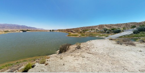 A serene landscape featuring a calm lake surrounded by dry hills and sparse vegetation under a clear blue sky.