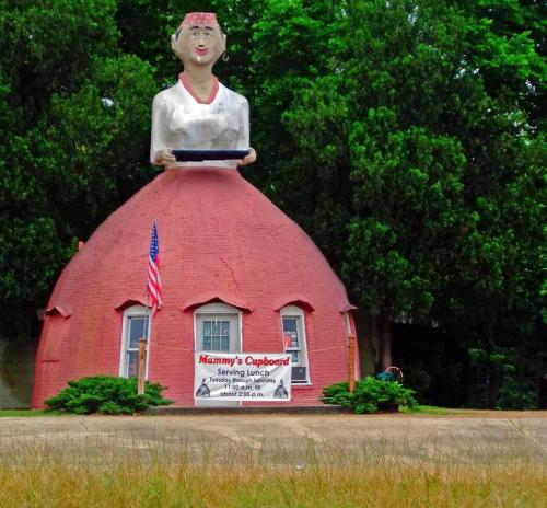 A quirky restaurant shaped like a large woman holding a tray, with a red dome structure and an American flag outside.