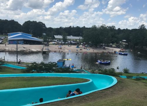 A water park scene with a winding slide, a sandy beach, and people enjoying the water and activities on a sunny day.