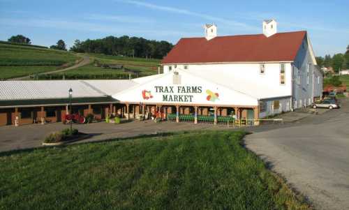 A large white building with a red roof, labeled "Trax Farms Market," surrounded by green fields and a clear blue sky.