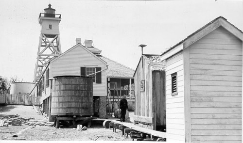 Historic lighthouse and buildings, with a person walking near a wooden barrel in a coastal setting.