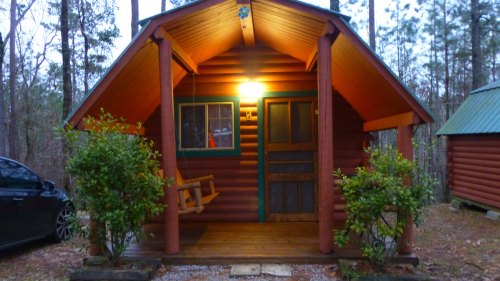 A cozy log cabin with a porch swing, surrounded by trees, illuminated by a warm light at dusk.