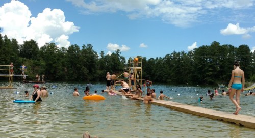 A lively swimming area with people enjoying the water, diving boards, and inflatable floats under a sunny sky.