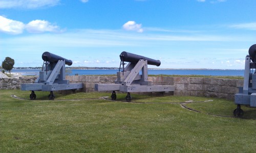 Three historic cannons on a grassy area overlooking a calm sea under a blue sky with scattered clouds.