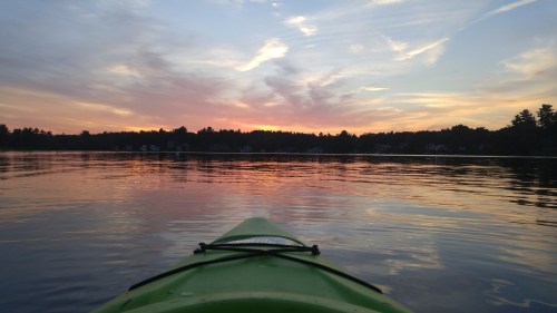 A green kayak on calm water at sunset, with colorful clouds reflecting on the surface and trees in the background.