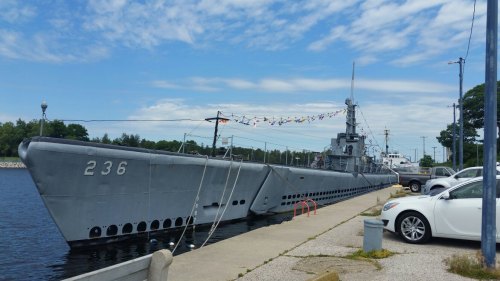 A gray submarine docked at a harbor, with colorful flags strung above and cars parked nearby under a blue sky.