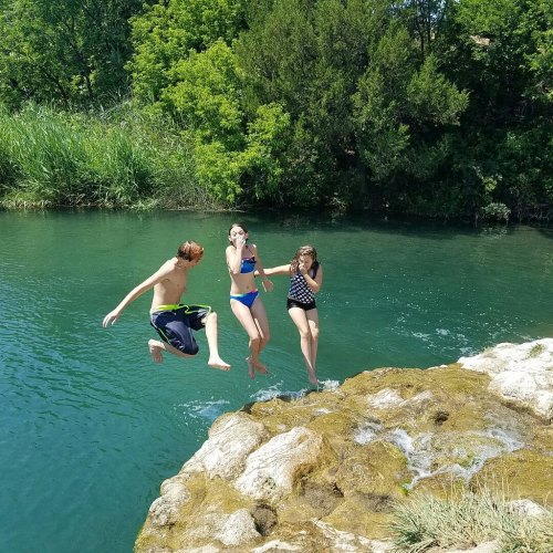 Three children jumping into a clear, green river from a rocky ledge on a sunny day, surrounded by lush greenery.
