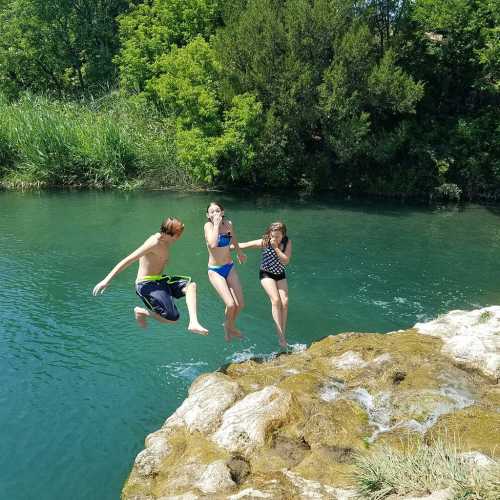 Three children jumping into a clear, green river from a rocky ledge on a sunny day, surrounded by lush greenery.