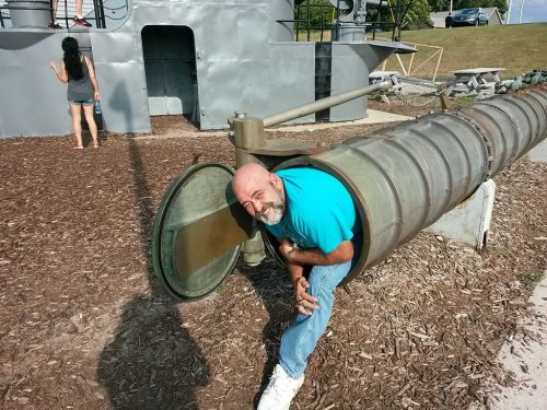 A man in a blue shirt smiles while emerging from a large metal pipe, with a woman in the background near a structure.