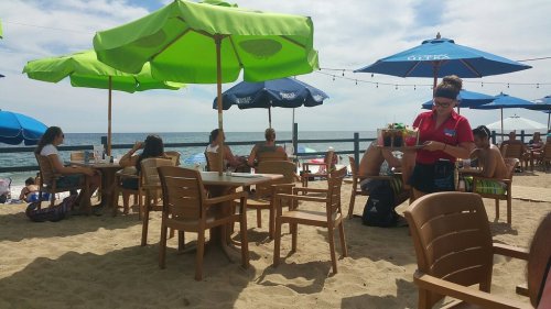A beachside café with colorful umbrellas, tables, and people enjoying the sun and ocean view.