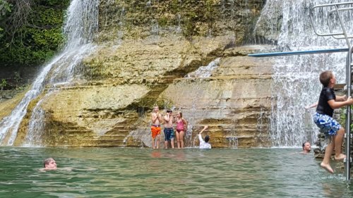 Children playing in a natural pool by a waterfall, some climbing a ladder, others swimming and enjoying the water.