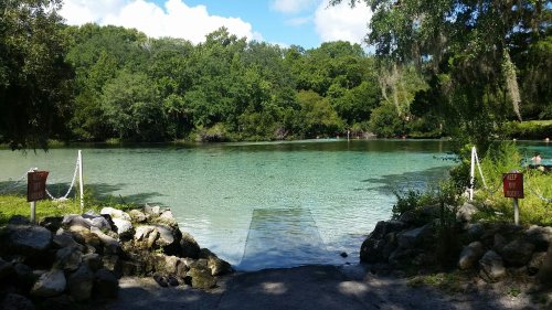 A serene lake surrounded by lush greenery, with a rocky launch area and clear blue water under a bright sky.