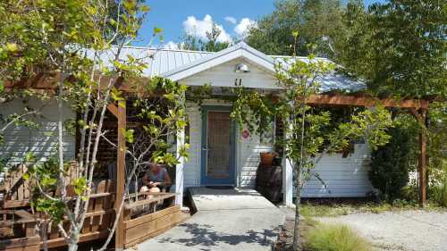 A small white house with a porch, surrounded by greenery and trees, under a blue sky with fluffy clouds.