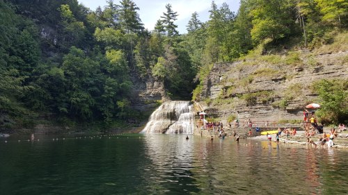 A scenic waterfall surrounded by lush greenery, with people swimming and relaxing by the water's edge.