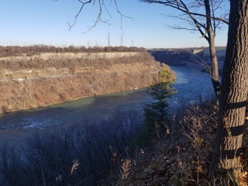 A scenic view of a river winding through a valley, flanked by trees and cliffs under a clear blue sky.