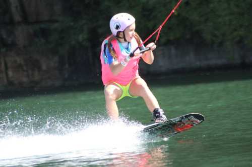 A young girl in a pink shirt and helmet enjoys wakeboarding on a calm lake, splashing water behind her.