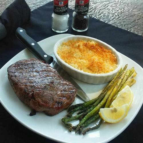 A plate featuring a grilled steak, asparagus, lemon slices, and a side of cheesy baked dish, with salt and pepper shakers.